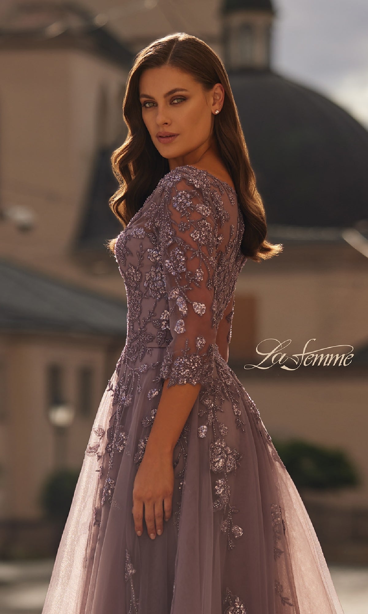 A woman stands outdoors, looking over her shoulder in the A-Line Tulle Sleeved Formal Dress: La Femme 32538, featuring a long lavender design with beaded lace appliques and tulle sleeves.